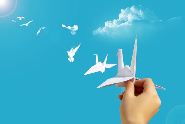 Hand holding an origami bird, leading to a trail of actual birds against a blue sky